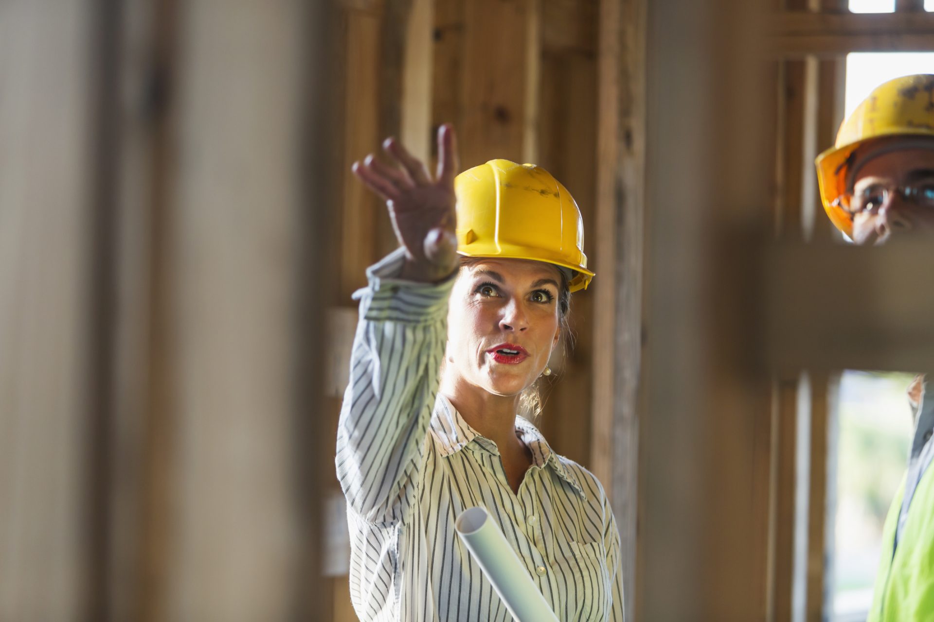Two workers talking at a construction site, standing in a building in the framing stage.  The focus is on the mature woman, wearing a yellow hard hat, holding a set of rolled-up plans, pointing upward.  The male worker is cropped and partly obsured by the wooden frame.