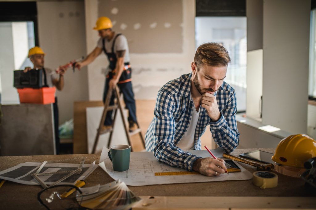 Pensive architect drawing improvements on housing project inside of a built structure. Workers are in the background.