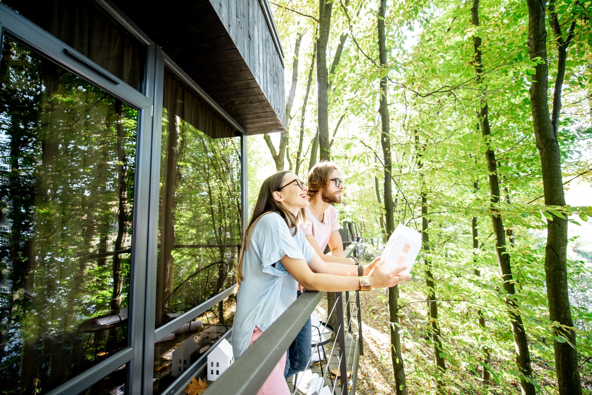 Young creative couple standing with house model on the balcony, designing or dreaming about eco houses in the forest