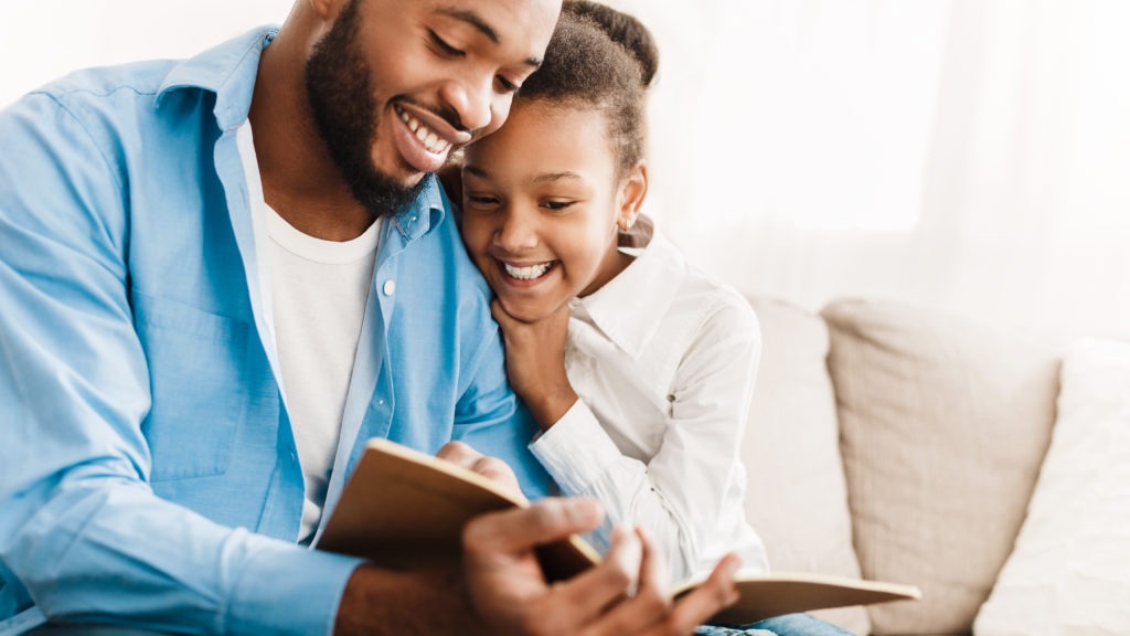 Father and daughter reading book together, sitting on sofa at home
