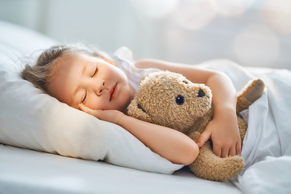 Adorable little child is sleeping in the bed with her toy. The girl is hugging the teddy bear.