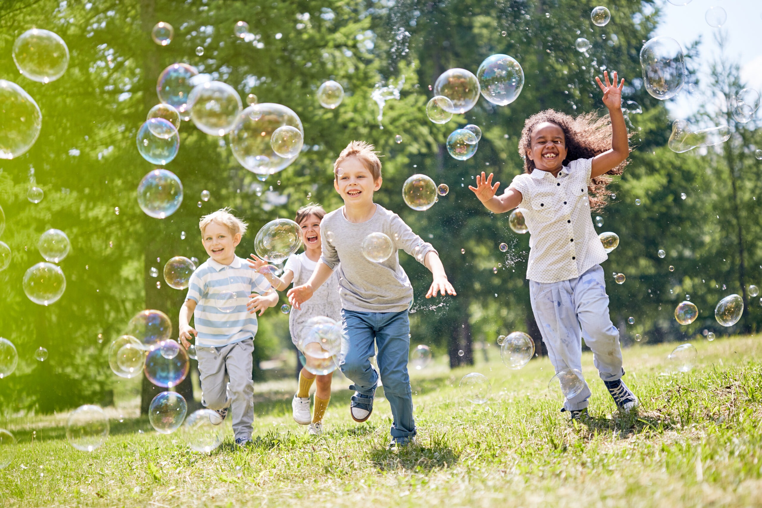 Multi-ethnic group of little friends with toothy smiles on their faces enjoying warm sunny day while participating in soap bubbles show