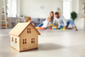 Un jeune couple avec enfant joue au fond de la pièce. au 1er plan, un jouet en bois représentant une maison