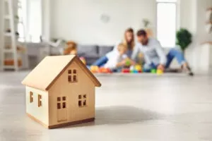 Un jeune couple avec enfant joue au fond de la pièce. au 1er plan, un jouet en bois représentant une maison